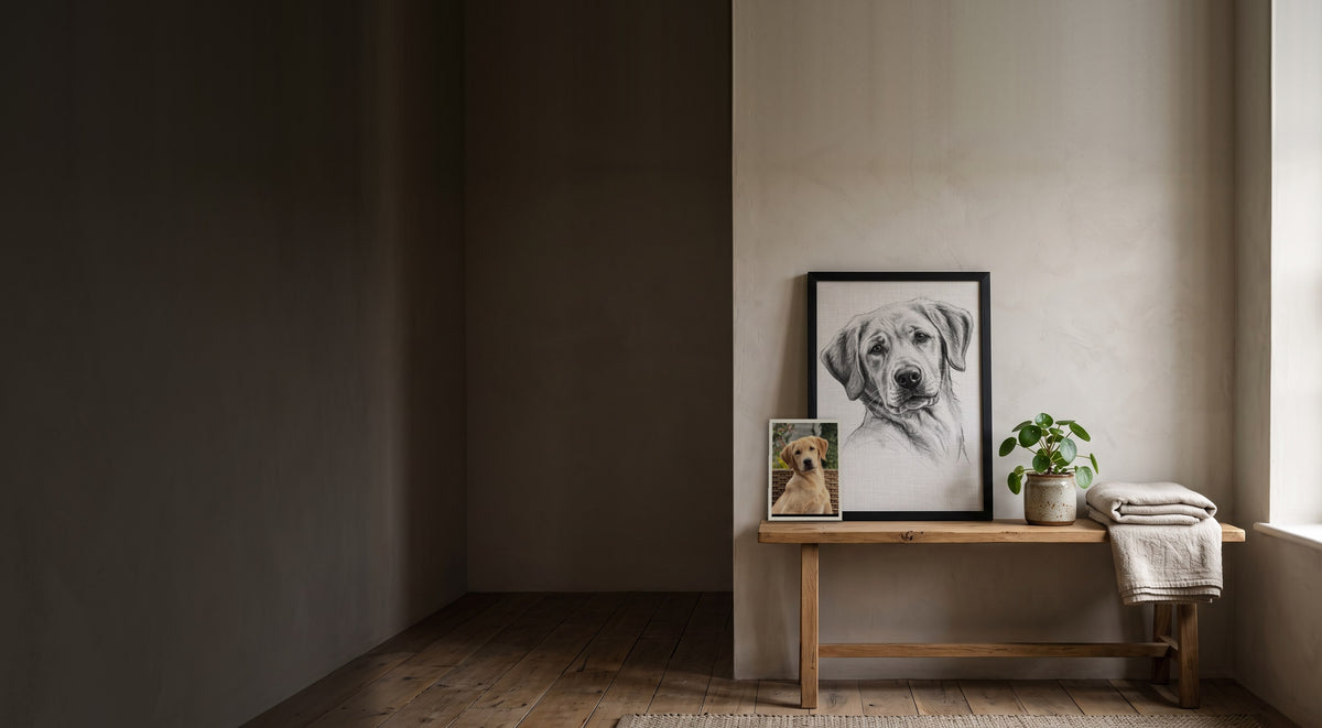 Charcoal portrait of a yellow Labrador in a black frame on an oak bench, with the original photo propped beside it